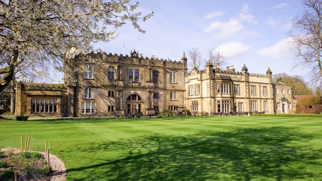 Historic stone building with manicured lawns, surrounded by trees and blooming flowers under a clear blue sky.