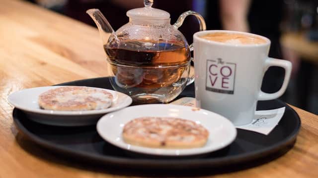 A tray with two cookies, a teapot, and a coffee cup bearing "The Cove" logo, set on a wooden counter.