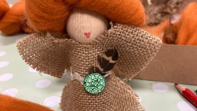 A handmade doll with orange hair, dressed in burlap, sits on a polka-dot table among crafting materials.