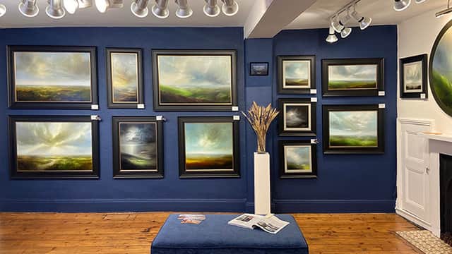 Gallery interior featuring framed landscape paintings on blue walls, wooden flooring, and a decorative plant display.