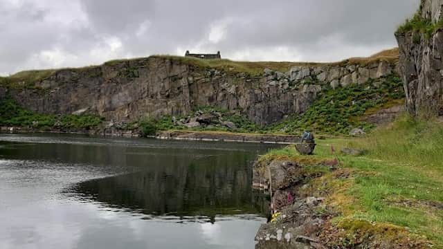 Serene waters reflect rugged cliffs under a cloudy sky, with a distant building atop the hill at Helwith Bridge.