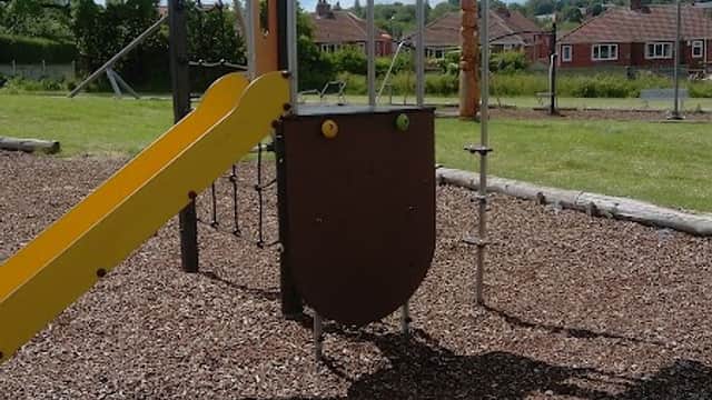 Colorful playground equipment with a slide, set on a gravel surface, surrounded by green grass and houses in the background.