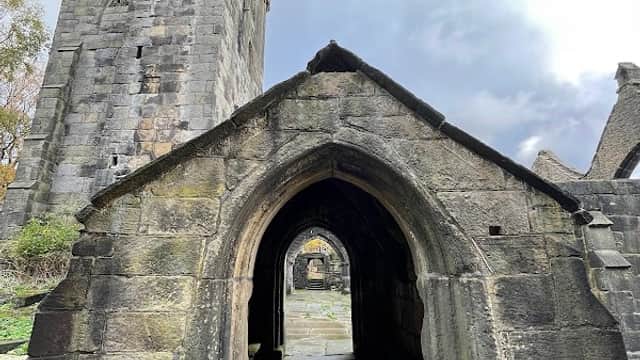 Archway leading to a stone structure with a tower, surrounded by greenery and a cloudy sky in Chiserley, Yorkshire.