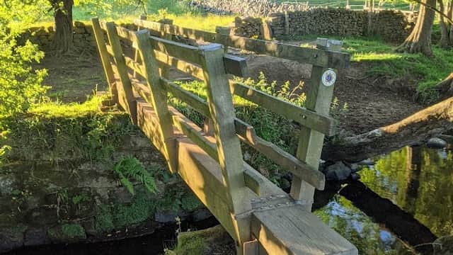 A wooden footbridge crosses a small stream, surrounded by lush greenery and trees in a tranquil campsite setting.