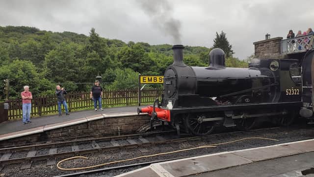 Steam locomotive at Embsay Railway Station with passengers observing and lush greenery in the background.
