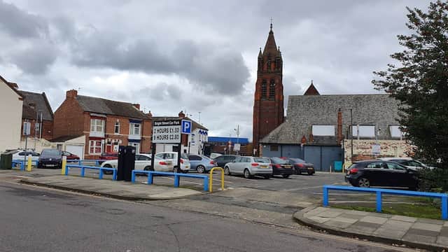 Pay & Display car park with parked cars, blue barriers, and a tall church tower in the background under a cloudy sky.