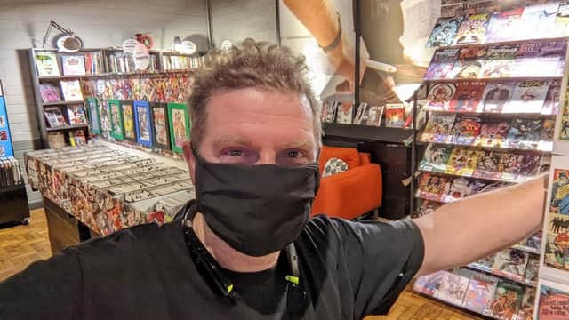 A man in a black mask stands in a vibrant bookshop filled with comics, colorful displays, and a cozy orange sofa.
