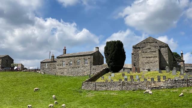 Stone buildings and a graveyard on a grassy hill, with sheep grazing under a partly cloudy sky in Keld, North Yorkshire.
