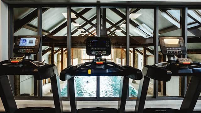 Treadmills with screens facing a swimming pool, framed by large windows and a wooden-beamed ceiling.