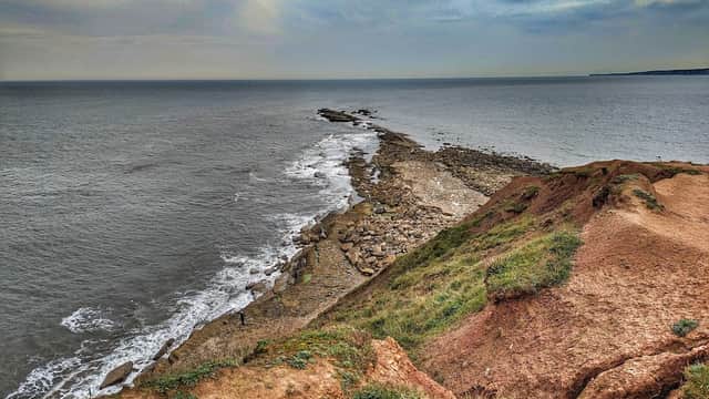 A rocky shoreline meets the sea under a cloudy sky, with grassy cliffs in the foreground.