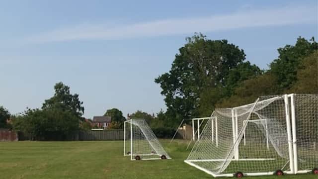 Football goals on a grassy field under a clear blue sky, surrounded by trees and distant buildings.