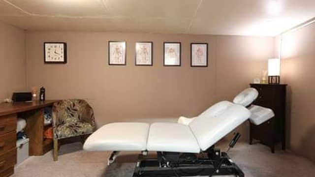 Treatment room with a white massage table, a desk, chair, and framed anatomical art on the walls. Soft lighting.