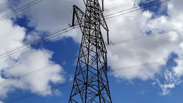 A tall power line tower against a backdrop of blue sky and fluffy white clouds.