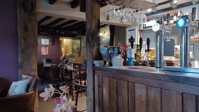 Cozy bar interior with wooden beams, a counter, hanging glassware, and seating areas in a rustic setting.