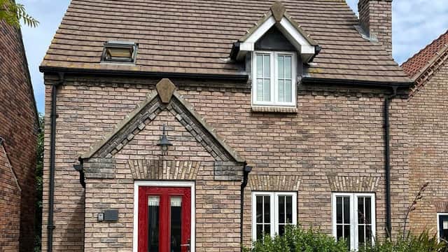 Brick cottage with a red front door, white windows, and a sloped roof, surrounded by greenery under a blue sky.