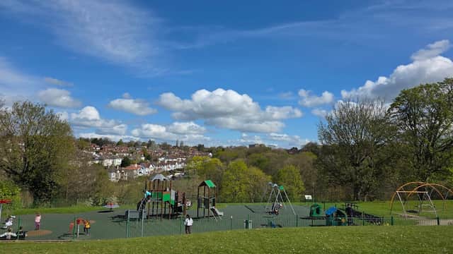 Colorful playground equipment in a park with green grass, trees, and a blue sky dotted with fluffy clouds.