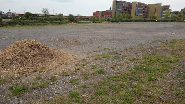Gravel car park with sparse grass, piles of mulch, and modern buildings in the background under a cloudy sky.