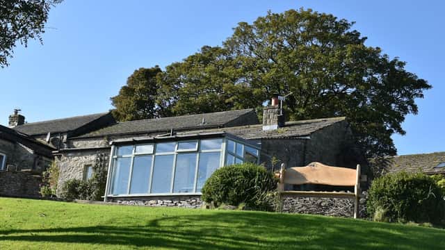 West Close Farmhouse features a stone exterior, large windows, and lush green surroundings under a clear blue sky.