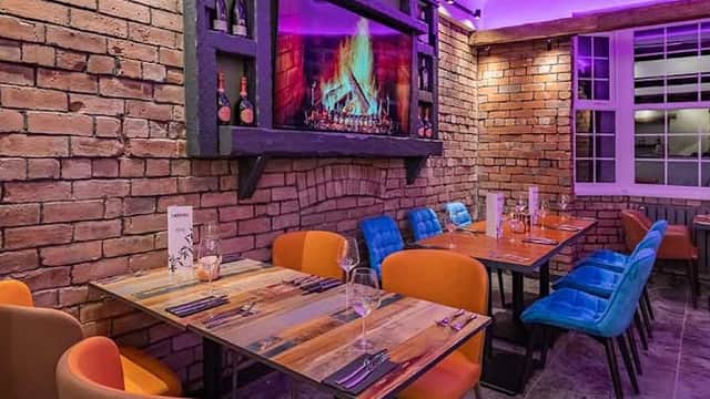 Cozy restaurant with colorful chairs, wooden tables, a brick wall, and a fireplace display under purple lighting.