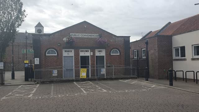 Public toilet building with three doors labeled for regulars, gentlemen, and ladies, surrounded by trees and parking.