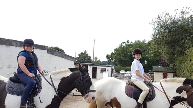 Two riders on horses at Follifoot Park Riding Centre, surrounded by trees and a stable building in the background.