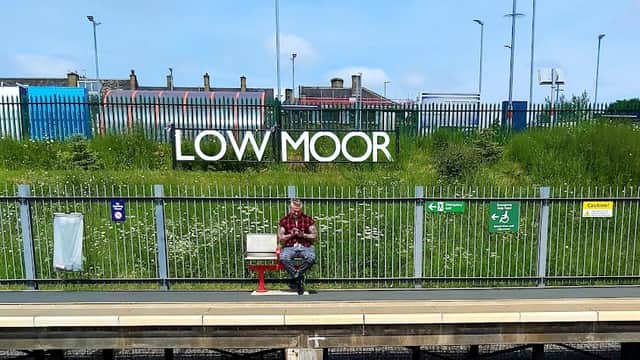 Low Moor station car park with a man sitting on a bench, greenery, and signage in the background.