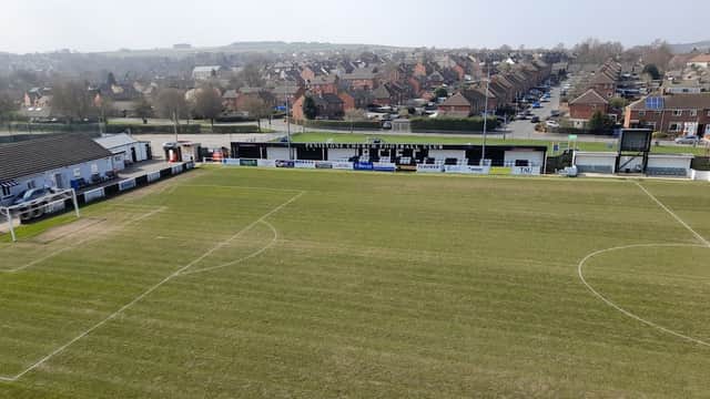 Green football pitch with stands, club signage, and residential area in the background under a clear sky.
