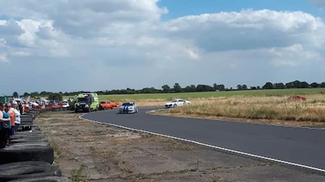 Go-karts racing on a track with spectators in the foreground and a blue sky with clouds above.