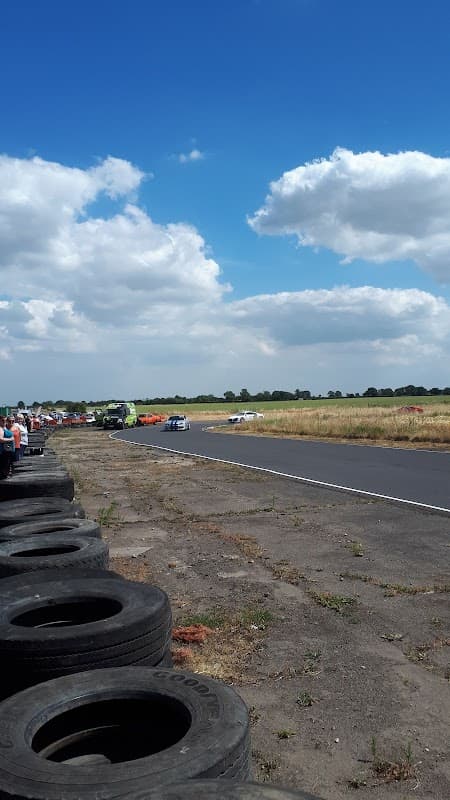 Go-karts racing on a track with spectators in the foreground and a blue sky with clouds above.