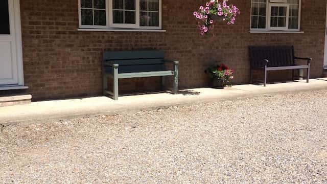 Single-story motel with a tiled roof, gravel pathway, benches, and flower pots in a sunny outdoor setting.