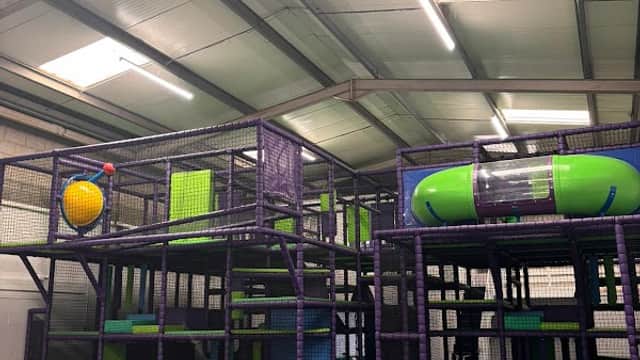 Colorful indoor soft play area with climbing frames, slides, and seating for kids and parents in a spacious setting.