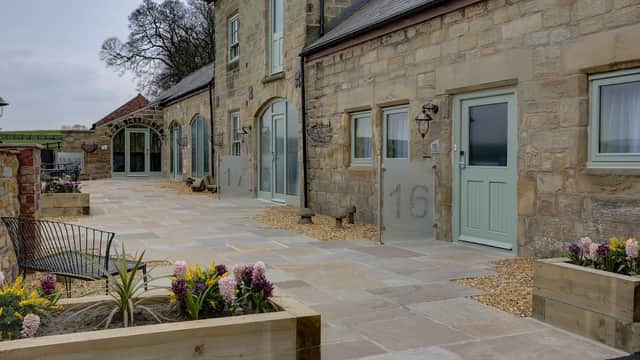 Historic stone building with large windows, numbered doors, and landscaped garden beds featuring flowers and seating.