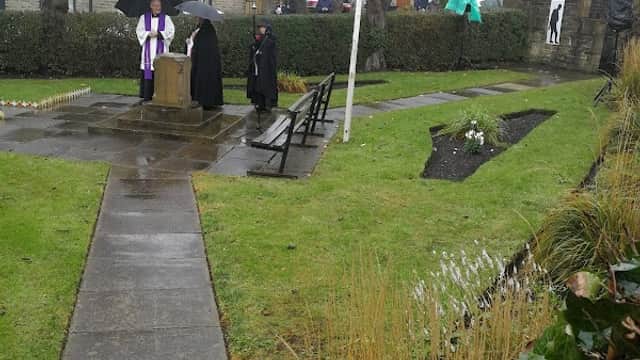 Memorial Gardens in Shelf, Yorkshire, featuring a stone monument, benches, and people under umbrellas in rainy weather.