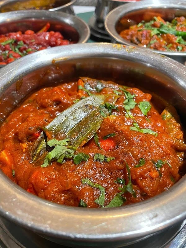 A rich, spicy curry topped with green herbs in a metal bowl, surrounded by other colorful dishes.