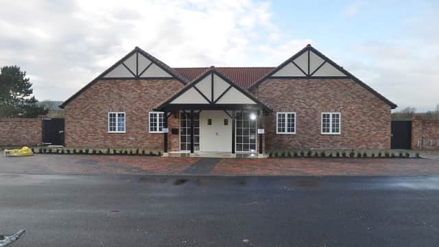 Brick building with a gabled roof, white windows, and a central entrance, surrounded by a paved area and greenery.