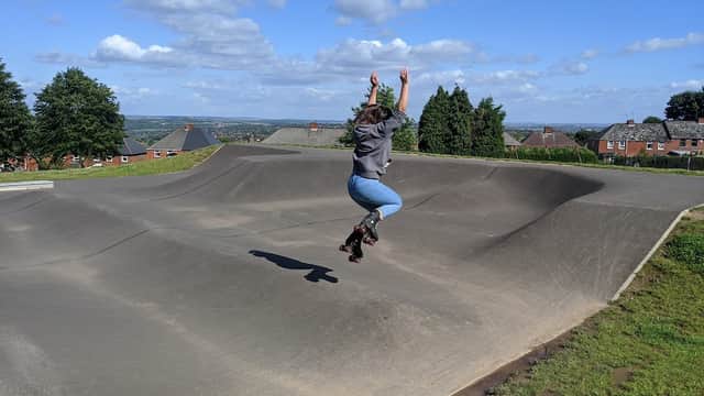 A person in roller skates jumps over a smooth, curved ramp in Grenoside Park, with houses and hills in the background.