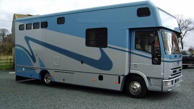 Blue and grey horsebox parked on gravel, with large windows and a streamlined design against a cloudy sky.