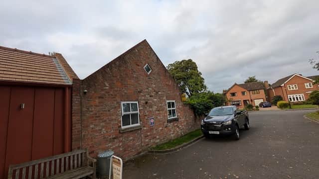 Brick building with a slanted roof, parked black vehicle, and residential homes in a quiet neighborhood.