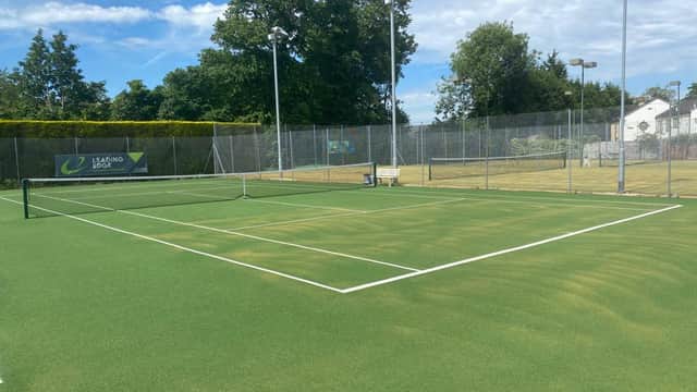 Green tennis court with a net, surrounded by trees and a clear blue sky at Liversedge Tennis Club.