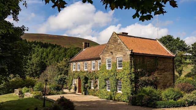 Stone country house covered in greenery, surrounded by gardens and hills under a partly cloudy sky.