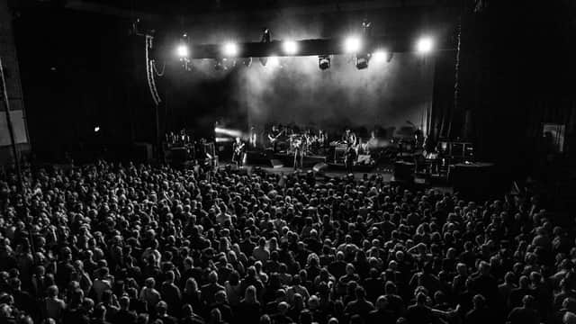 Crowded concert hall with band performing on stage under bright lights, audience seated and standing in darkened space.