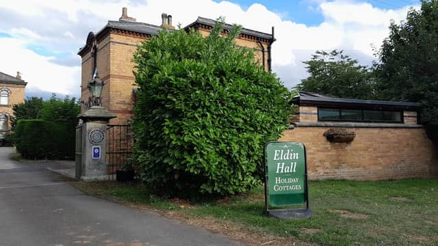 Eldin Hall Cottages sign near a lush green bush and brick building, with a cloudy sky in the background.