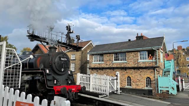 Steam train passing through Grosmont Car & Coach Park, with a historic building and cloudy sky in the background.