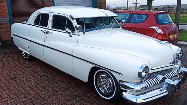 Classic white car parked outside a garage, with a red vehicle nearby and a brick wall in the background.