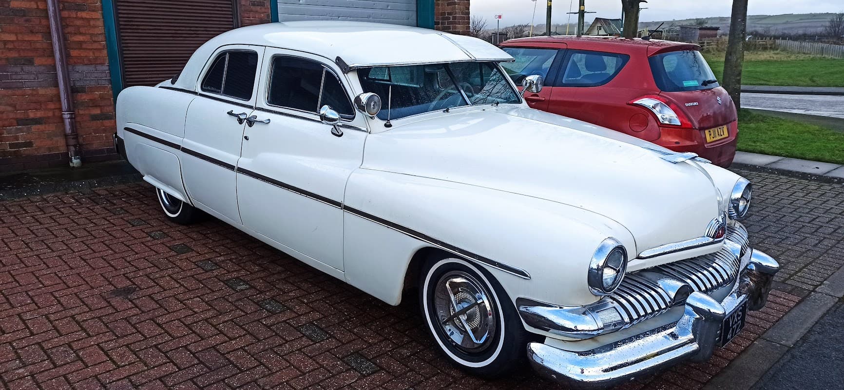 Classic white car parked outside a garage, with a red vehicle nearby and a brick wall in the background.
