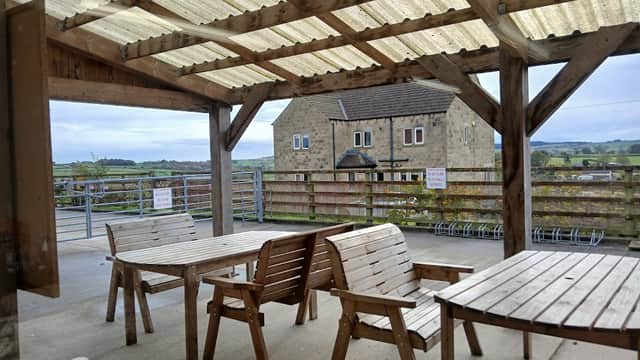 Wooden tables and benches under a covered patio, with a stone building and green hills in the background.