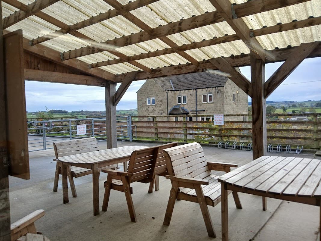 Wooden tables and benches under a covered patio, with a stone building and green hills in the background.