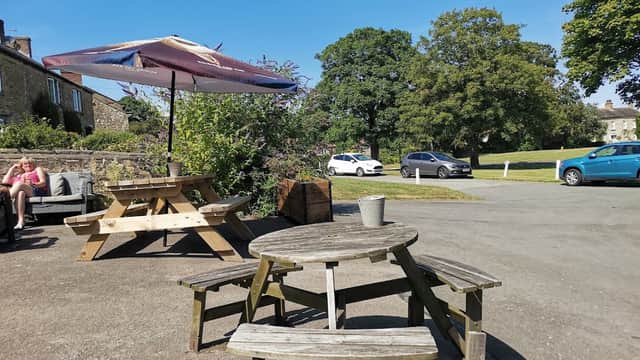 Outdoor seating area with wooden tables and umbrellas, surrounded by greenery and a clear blue sky in Crakehall, Yorkshire.