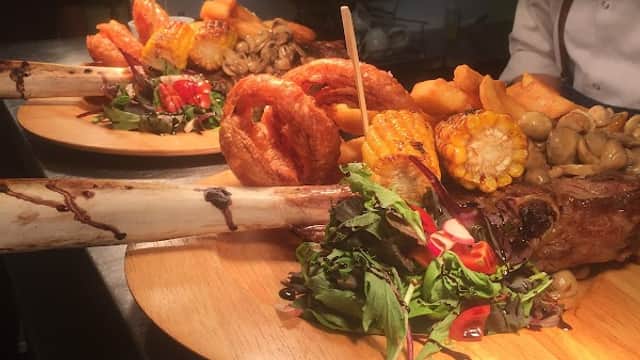 Two beautifully plated dishes featuring grilled meat, roasted vegetables, onion rings, and fresh greens on wooden boards.