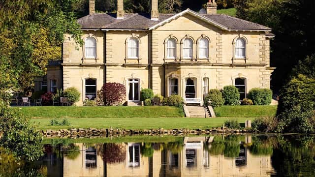 Elegant stone mansion with large windows, surrounded by lush greenery and reflecting in a calm lake.